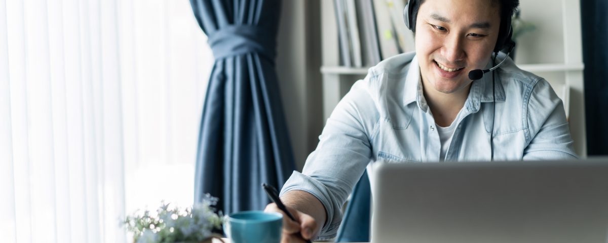 Asian businessman talking to colleague team in video call conference writing note on book with smile face. Man using computer laptop and headphone for online meeting. Smart working from home concept.