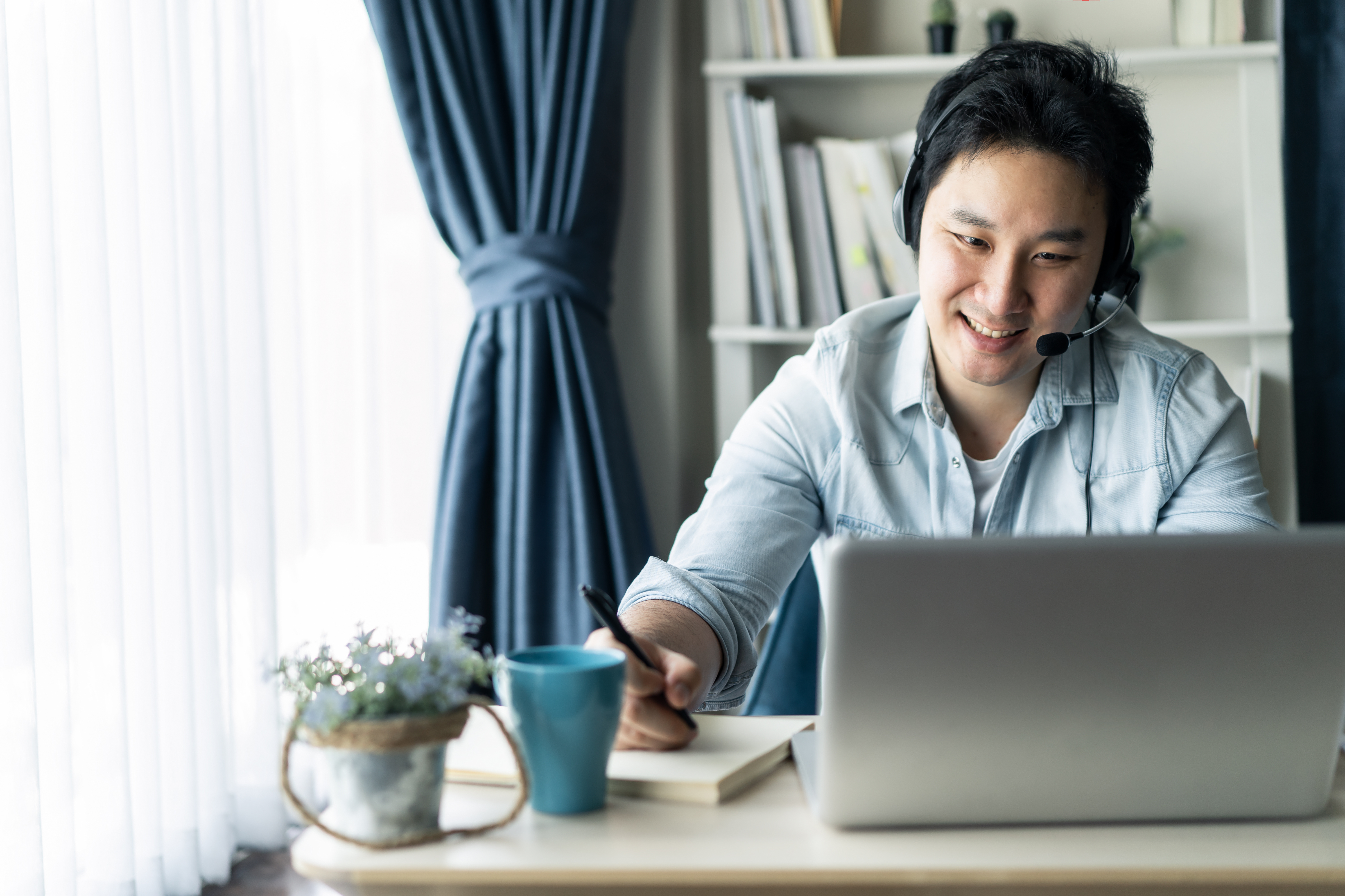 Asian businessman talking to colleague team in video call conference writing note on book with smile face. Man using computer laptop and headphone for online meeting. Smart working from home concept.